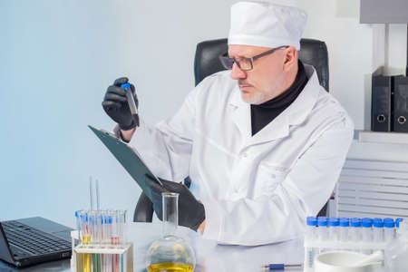 A doctor in a white coat and cap examines the substance in a plastic test tube. The person compares the substance in the flask with the test values. Medical test. Medical diagnostics.の写真素材