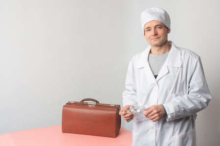 A doctor in a white coat with a medical bag. The doctor is holding a set of individual bottles for disinfectants. Portrait of a medic on a white background. A calm doctor with a set of containers.の写真素材