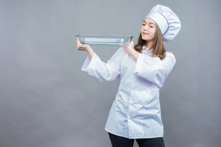 A young woman works as a Baker. The pleasure of cooking. Portrait of a young girl-chef. The girl is holding a glass baking dish. A Baker in an apron and cap on a gray background.の写真素材