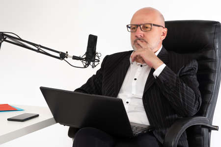 Remote work of a businessman. A thoughtful man with a laptop and a microphone. A man in a business suit sits with a laptop on his lap. A businessman is sitting at a table with a condenser microphone.の写真素材