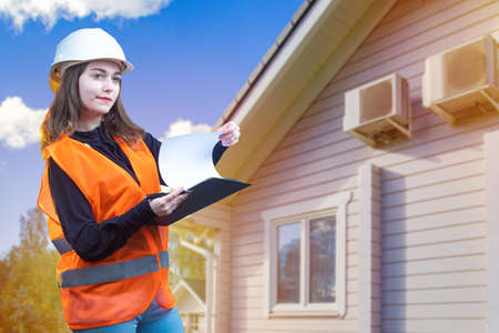Construction engineer. Acceptance of construction works. The girl works as a civil engineer. Female architect on the background of a cottage. A girl in a white helmet and orange vest.の写真素材