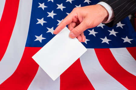 US President Election. An American is voting in the 2020 election. A man's hand with a ballot on the background of the American flag. A person at a polling station in the United States.の写真素材