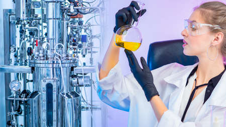 Girl with a flask on the background of a laboratory bioreactor. Biochemistry. Research in the field of pharmacology. The reaction of fermentation. Laboratory equipment.の写真素材