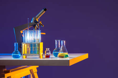 Table with a microscope and test tubes. Flasks with multicolored chemical liquids and a microscope on a lilac background. Chemical concept with space for text. Brightly lit chemical laboratory.の写真素材