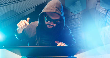 The concept of cybercrime. Hacker on the background of skyscrapers. A man with a closed face at the computer. Crimes on the Internet. A man in black glasses and a hood writes a computer program.の写真素材