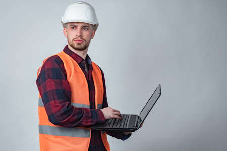 A man in an orange vest and hard hat is holding a laptop. Construction engineer. The architect in the hard hat looks away. Monitoring the progress of construction. Checking all stages of construction.の写真素材