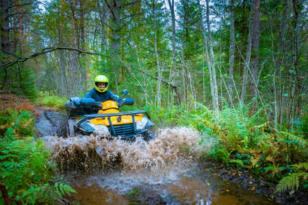A man rushes on an ATV on a forest road. Yellow ATV drives through a puddle on a forest road. Cross-country racing. Quad bike racing. Extreme ATV riding. Rider on a Quad bike in the forest.の写真素材