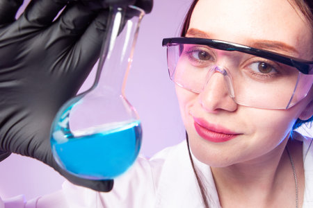 The face of a chemist girl in safety glasses. A woman is holding a flask with a blue chemical...