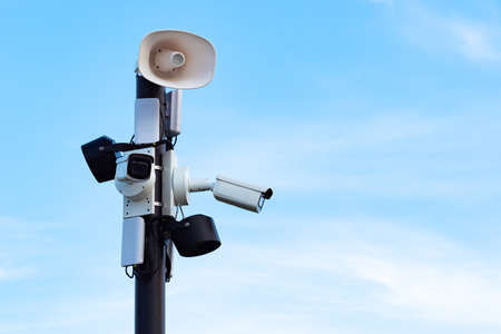 Security cameras and speakers on a pole above a blue sky. Close-up of outdoor surveillance cameras, floodlights and loudspeakers on a street pole. Ensuring security on the street.の写真素材