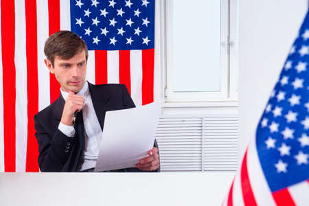 A man studies documents against the background of American flags. Work in the USA. Employment in America. A man is looking for a job in the United States of America. Moving to the United States.の写真素材