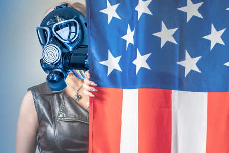 A girl in a gas mask stands behind an American flag. A woman wearing a respirator and a leather vest next to the flag of the United States. Emergency respirators in the United States.の写真素材