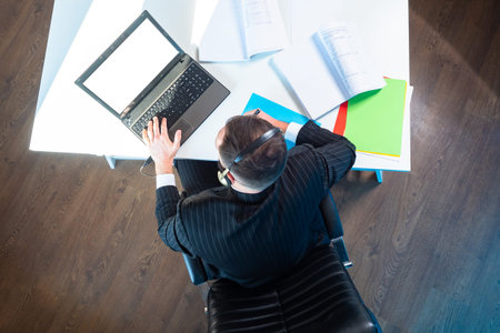 A person working remotely is photographed from above. The man looks at his laptop and makes notes on...