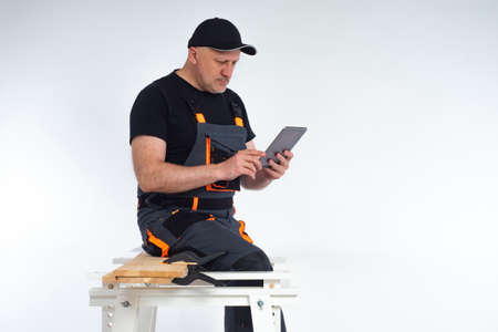 A worker sits on a workbench and looks at a tablet computer. Master with an electronic tablet in his hands. The carpenter specifies the details of the project via the Internet.の写真素材
