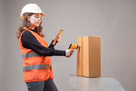 Warehouse storage. A girl with a barcode reader and a tablet in her hands. A girl in a hard hat and orange vest reads a barcode from a box.の写真素材