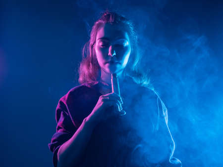 VAPE Smoking. A girl with closed eyes smokes a VAPE. The pleasure of Smoking an electronic cigarette. VAPE shop. Gadgets for Smoking. The concept of Smoking an e-cigarette in blue.の写真素材