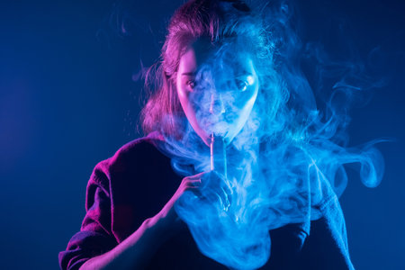 Smoking an electronic cigarette. The girl's face is covered with smoke from vape. Portrait of a female smoker in a cloud of smoke. A woman smokes a VAPE on a dark background.の写真素材