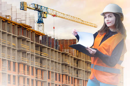 The girl works as a construction engineer. A girl in a white helmet on the background of a building under construction. A woman in an orange vest against the background of a crane.の写真素材