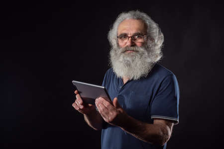 A gray-haired man with a beard holds a tablet in his hands. An elderly man with glasses on a dark background. An intelligent old man with a tablet in his hands.の写真素材