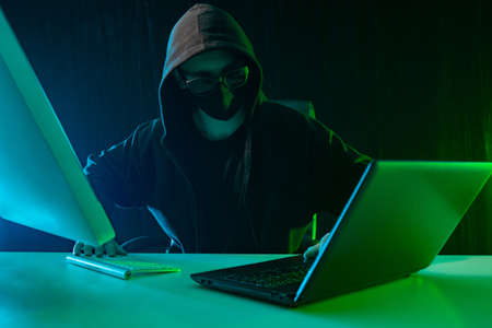 Cybercrime. Covered face man behind computers. Committing a crime using a computer. The concept of cybercrime against a dark background. Hacking computer programs.の写真素材