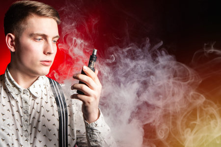 The concept of vaping in red. A young man with an electronic cigarette in his hands against the background of smoke. Space for text. VAPE shop. Smoking electronic cigarettes. The pleasure of vaping.の写真素材
