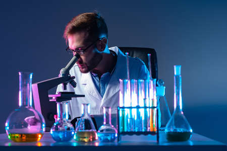 Microbiologist at work. A man looks through a microscope against the background of test tubes. A scientist makes scientific experiments. Work in a research laboratory.の写真素材