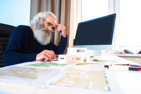 A man with gray hair and a beard looks thoughtfully at the model of the house. Work on a...