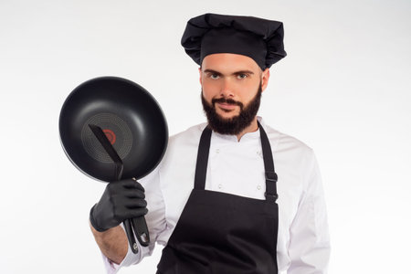 The bearded chef smiles at the camera. A man in a chef uniform holds a frying pan in his hand....
