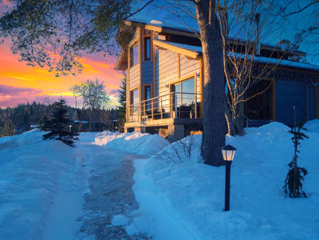 Sunset sky and country house. Cottage on the background of a sunset in winter. Private house with a large balcony. Winter outside the city. Paths in the snow lead to the cottage.の写真素材