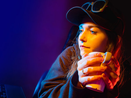 Portrait of a girl with a mug in her hands. A long-haired girl in a black baseball cap. The beautifully lit face of a young woman. Pensive girl on a dark background.の写真素材