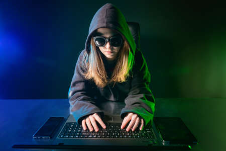 The girl hacker. Cybercrime. The girl breaks into a computer program. The girl at the computer hides her face. Violation of law. The person hides his face behind a hood and black glasses.の写真素材