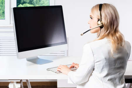 A girl in a white suit and headphones. A woman works at a computer. Telecommuting. The blonde consults clients through a computer application. A beautiful girl is sitting at her Desk.の写真素材