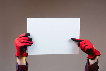 The person holds a white sheet over his head and points at it. Space for text. Attracting attention to the ad. Hands in red construction gloves with a poster without text.の写真素材