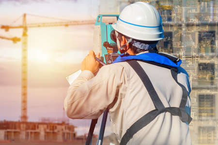 A surveyor works on a construction site. Measuring equipment in construction. A man looks out of the window of the theodolite. A man in a white construction helmet works with a geodesic tool.の写真素材