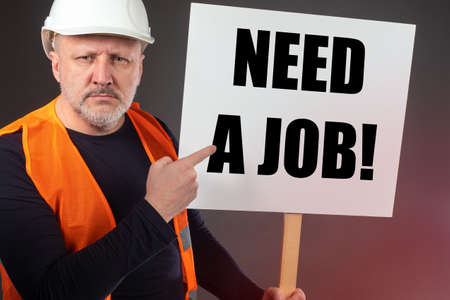 A man in a construction helmet points to a poster about job search. Unemployment. The loss of a job due to the coronavirus. Ruin of construction companies. Deprivation of means of livelihood.の写真素材