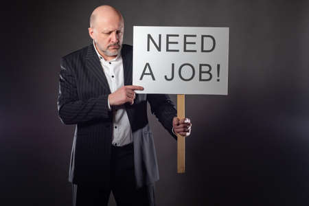 A sad man in a suit with a poster needs a job. Unemployment. Job loss. Deprivation of means of livelihood. Lack of jobs. Plant closures. Crisis. Collapse. Fear of the future.の写真素材
