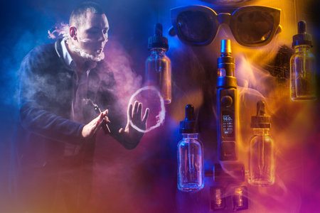The man lets out smoke from his mouth and nose in an unusual shape. The concept of vaping. A man with a VAPE gestures against the background of gadgets for smoking. Fashion for Smoking e-cigarettes.の写真素材