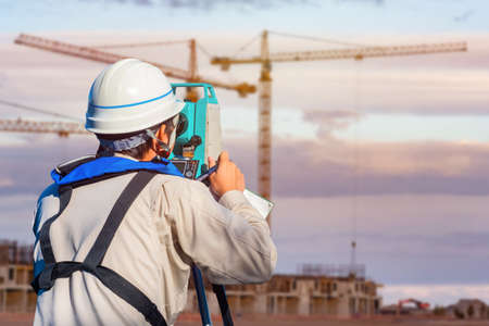 Surveyor on a construction site. A man takes measurements on a construction site. Geodesy and cartography. Measuring equipment. Builder, theodolite, and houses under construction.の写真素材