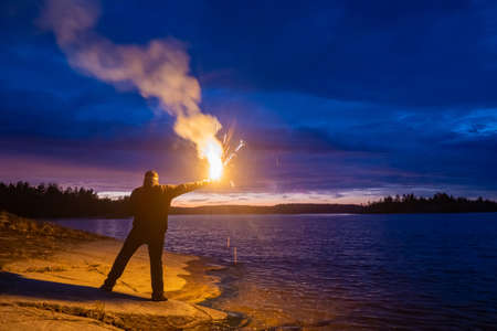 Smoke signal. A man with a smoke torch in the evening by the water. Rescue. SOS. Distress signal. The man lights a smoke signal and calls for help. A man in a life jacket on the lake shore.の写真素材