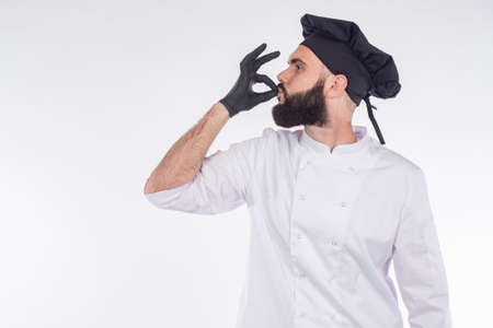The chef approves. The bearded chef makes a sign with his hands very tasty. Excellent cuisine. A chef in a black hat and gloves. Man on a white background with a space for text.の写真素材