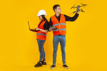 Drone in hands of builders. Guy and a girl launch a drone. Concept - shooting services with a quad copter. Workers in orange vests are filming from a drone. Guy and girl on orange background. Laptopの写真素材