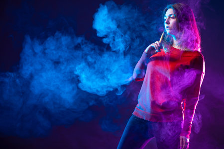 Young woman is wapit. Girl in a red blouse smokes a vape. Student holds a cigarette to her mouth....
