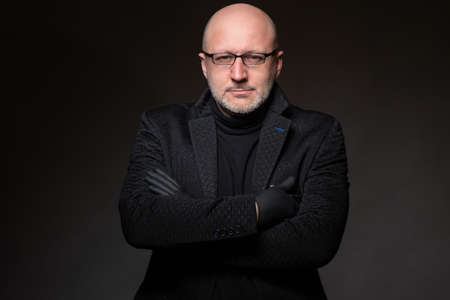 Adult human on a dark background. A man with glasses is looking at the camera. Portrait. Bald man in dark clothes. Concept - human in mourning clothes. Adult man in dark overcoatの写真素材