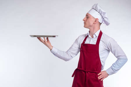 Chef is holding an empty tray. Man chef with a metal toe cap in his hands. Chef on a white background. Concept - he demonstrates a dish. The cook shows something. Man in cook uniform.の写真素材