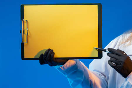 Yellow sheet of paper in clipboard. Man in a white coat shows a blank sheet of paper. Concept - it indicates some kind of signature. Place for inscription on clipboard of doctor. Blue backgroundの写真素材