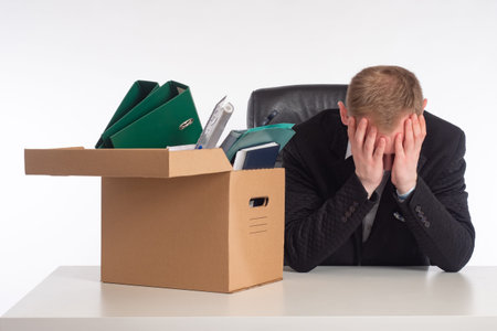 Cardboard box in front of man. Box as symbol of dismissal. He is upset by his dismissal. Fired man covers his face with his hands. Dismissed employee sits at table. Dismissal man on white backgroundの写真素材