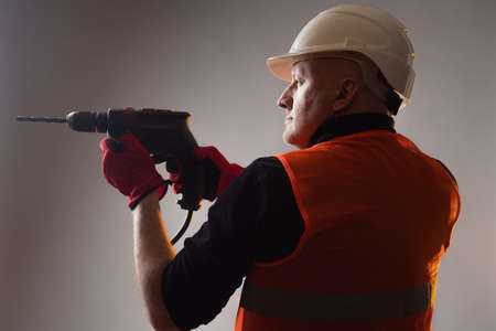 Builder holds a drill as a weapon. Builder works with an electric drill. Concept - human wants to drill a hole. Concept - sale of equipment for repair work. Construction works. Man in an orange vestの写真素材