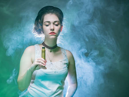 The girl with the VAPE is illuminated from behind. The woman holds a VAPE in her hand and looks down. A girl in a cloud of smoke from an electronic cigarette. The concept of vaping, vapeshop.の写真素材