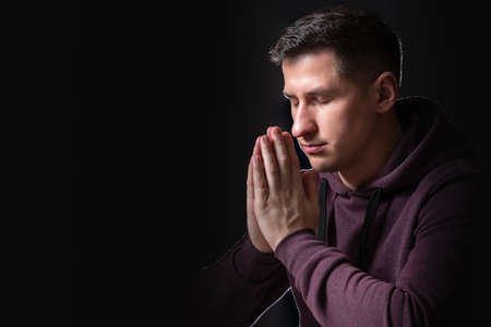 A young man praying. The person has folded his hands in front of him, closed his eyes, and communicates with God. Prayer of a young man. A religious man on a black background.の写真素材