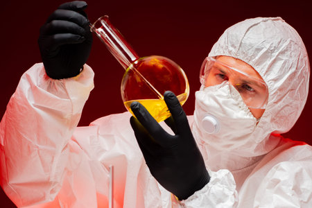 A chemist works with dangerous substances. A man in protective overalls examines a flask with a...