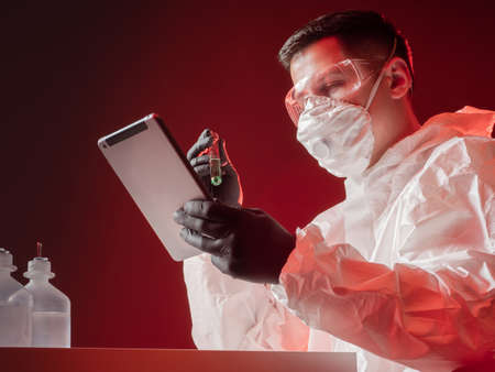 Laboratory analysis. A chemist with a test tube and a tablet computer in his hands. A man in a protective suit works in a chemical laboratory.の写真素材